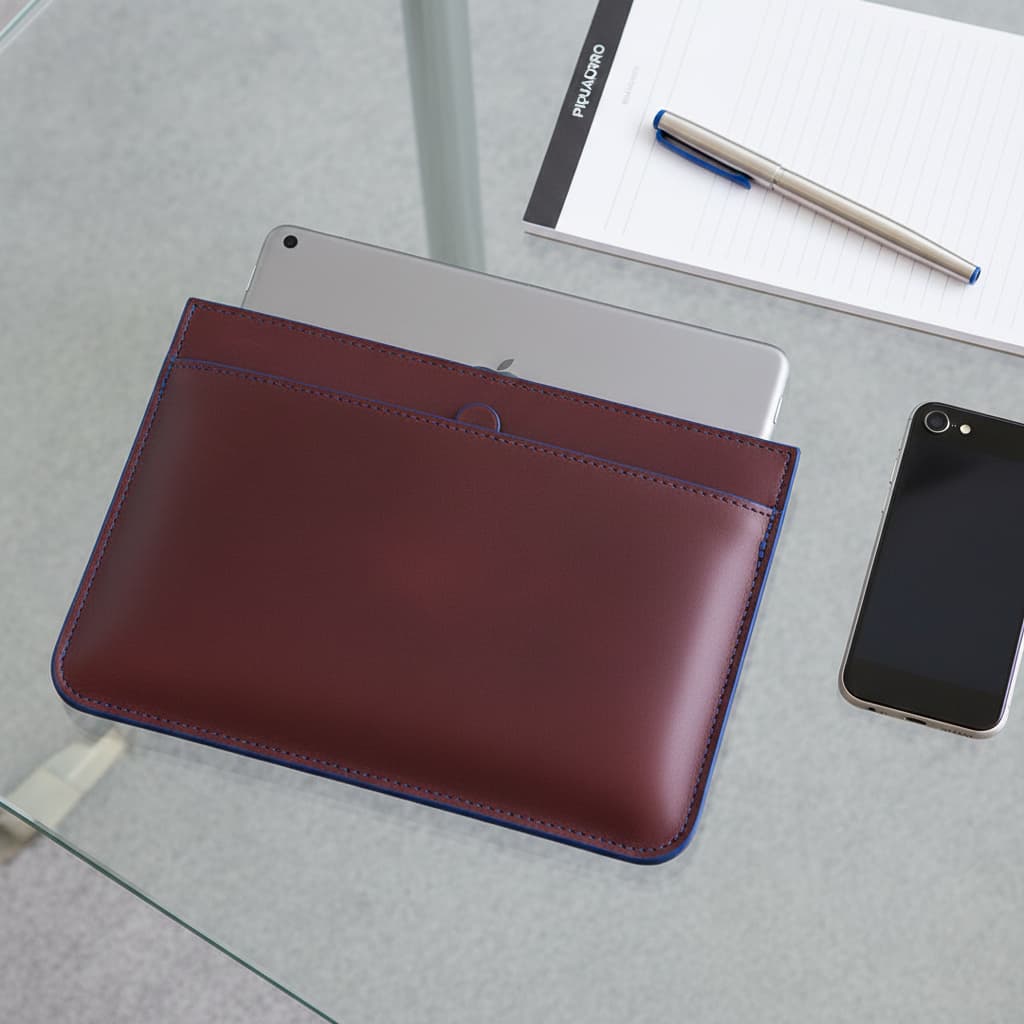 Sacoche pour iPad Piquadro Blue Square en cuir acajou avec bordures bleues posée sur un bureau en verre, un iPad Pro partiellement inséré, accompagnée d'un stylo et d'un smartphone.