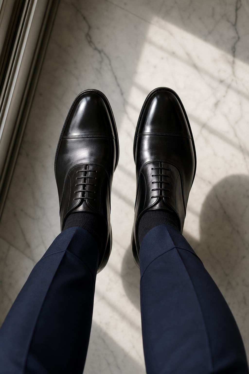 Vue de dessus de chaussures oxford noires cirées avec laçage droit, portées avec un pantalon bleu marine, posées sur un sol en marbre, éclairées par la lumière naturelle.