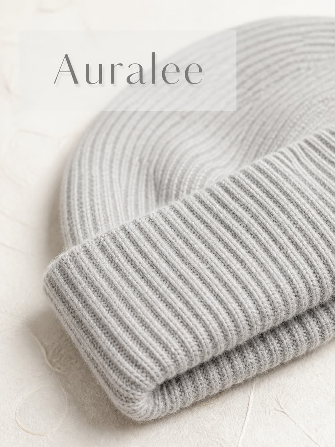Vue macro d'un bonnet Auralee en cachemire bébé gris pâle, mettant en valeur ses fibres délicates et sa texture douce, sur fond de papier washi japonais, avec un éclairage diffus et une surimpression du texte