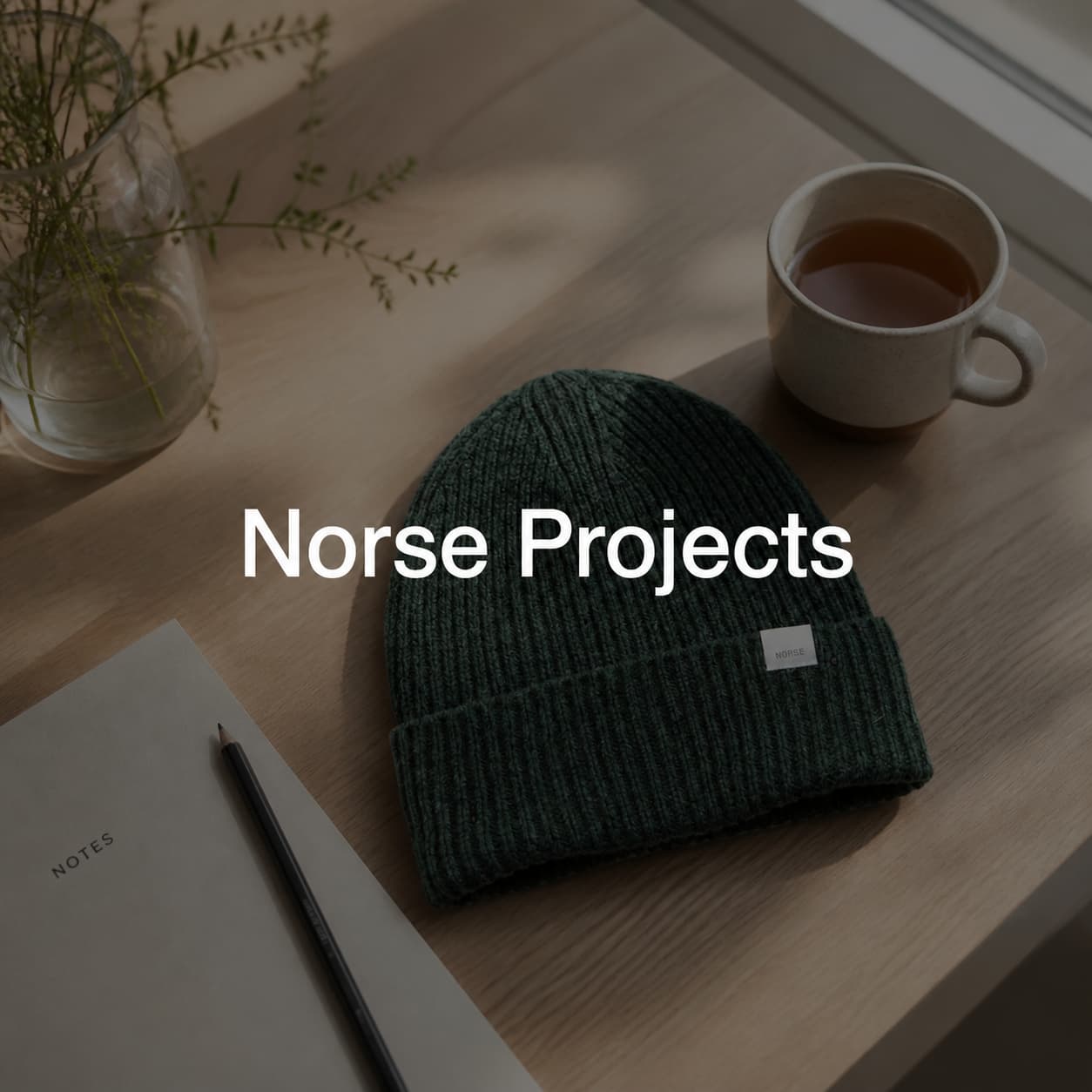 Bonnet en laine mérinos vert forêt de la marque Norse Projects posé sur une table en bois à côté d'une tasse de thé et d'un carnet, dans une composition minimaliste de style scandinave.
