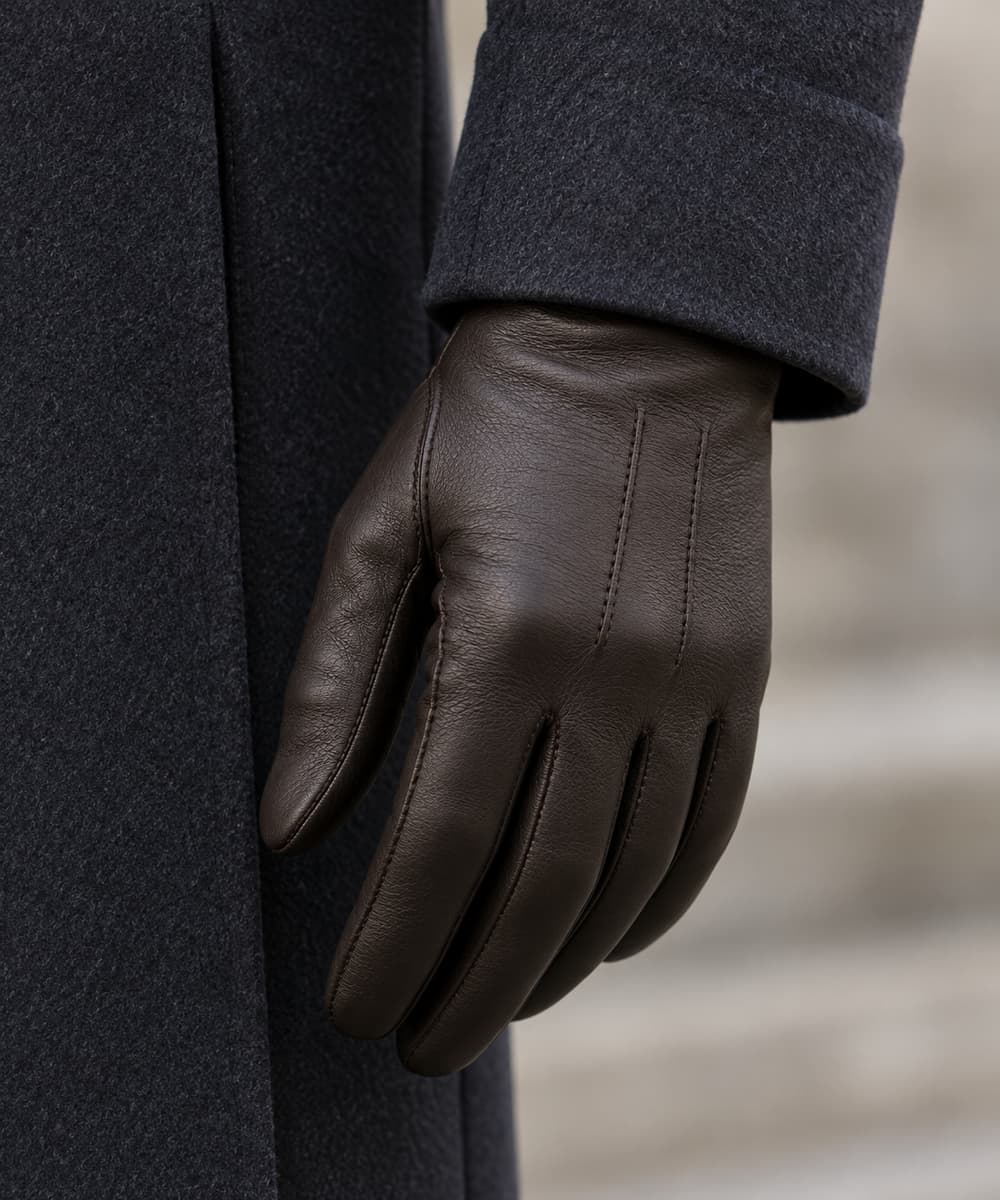 Main portant un gant en cuir lisse sombre au bout d’une manche de manteau en laine, avec des coutures discrètes et une coupe ajustée.